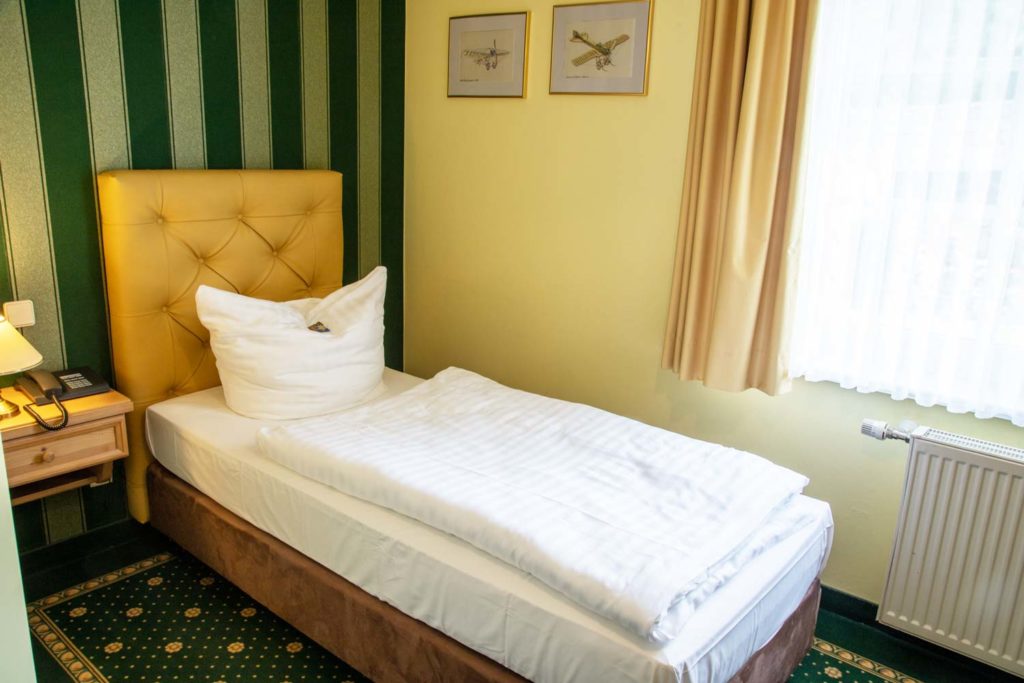 Das Foto zeigt ein gemütliches und komfortables Einzelbett mit weicher, hoher Matratze und frischem weißen Bettbezug in einem Raum mit gediegener Tapete und historischem Musterteppich im Hotel Fliegerheim in Borkheide.