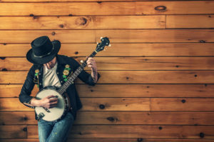 Das Foto zeigt einen Mann mit Cowboyhut der auf einer banjogitarre spielt und dient als Beitragsbild zur musikalischen Veranstaltung Sunday Lunch im Hotel Restaurant Fliegerheim in Borkheide bei Beelitz in Brandenburg.